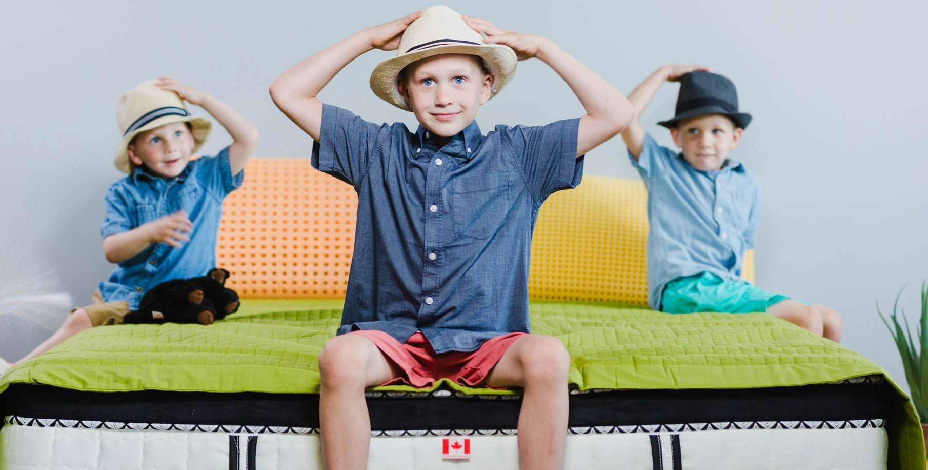 Three children sitting on a mattress with colorful covers and hats, in a room setting.